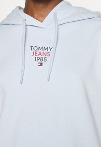 Sweat à capuche bleu clair avec le texte "TOMMY JEANS 1985" et un petit logo rouge, blanc et noir sur la poitrine, porté par une personne.