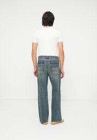 Heren blauwe denim jeans met een relaxte pasvorm, voorzien van brede pijpen, achterzakken en contrasterende stiksels. Gecombineerd met een effen wit T-shirt.