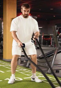 Uomo in una camicia bianca a maniche corte e pantaloni corti si allena con delle corde da battaglia nere su un pavimento sportivo sintetico verde. Equipaggiamento da ginnastica visibile sullo sfondo.