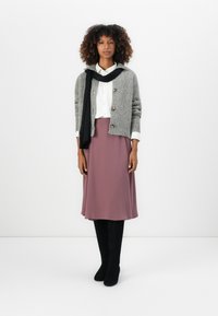 Cardigan grigio a maglia con bottoni, camicia bianca, gonna rosa pastello e stivali neri al ginocchio. Sciarpa appoggiata attorno al collo, il tutto su uno sfondo semplice.