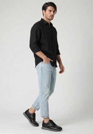 Muž stojí a má na sobě černé tričko s dlouhým rukávem, světle modré džíny a černé tenisky na jednoduchém světlém pozadí.