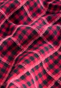 Sarkans un melns šķērsotais audums ar mīkstu, teksturētu virsmu, kurā ir izteikti kvadrātu raksti un kontrastējošas šuvju līnijas.