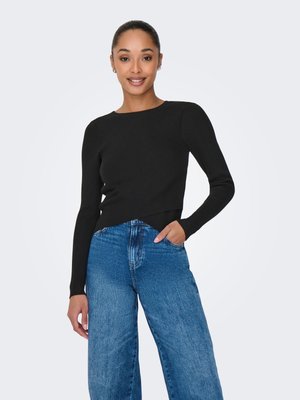 Kvinne med håret trukket bakover iført en svart ribbet genser med lange ermer og løse blå jeans, smilende med en hånd i lomma.