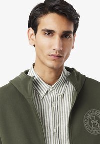 Felpa verde con zip sopra una camicia a bottoni bianca e verde a righe. Presenta una patch con il logo e un tessuto testurizzato. Vista ravvicinata del colletto.