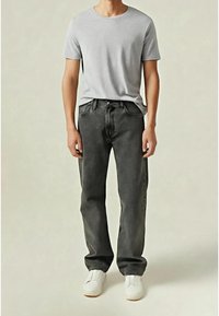 Personne portant une chemise à manches courtes gris clair, un jean gris foncé et des chaussures blanches sans lacets, debout devant un fond uni.