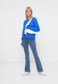 Cardigan en maille bleue avec un col rond, un top blanc en dessous, un jean bleu à motifs évasés et des baskets blanches. Design simple et épuré.