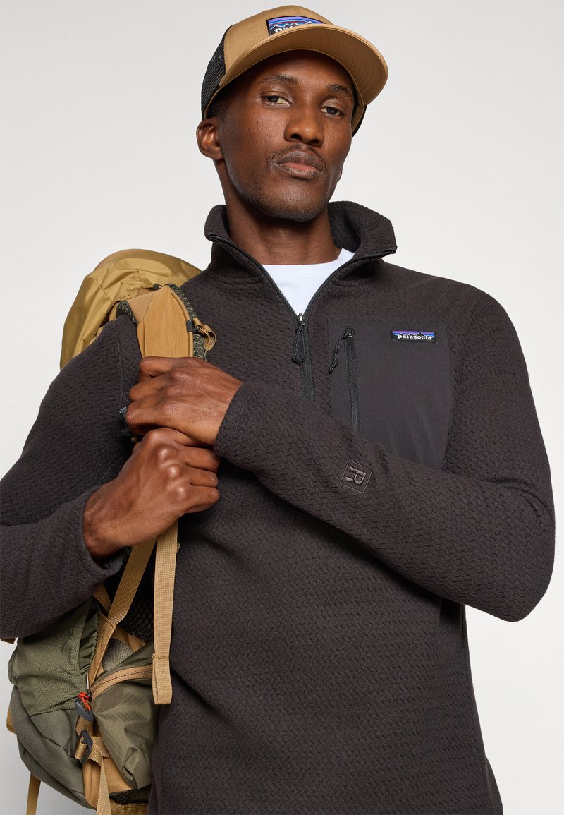 Schwarzer Fleece-Pullover mit strukturiertem Muster, halber Reißverschluss vorne und Brusttasche. Beigefarbene Kappe mit Netzrücken. Grüner und sandfarbener Rucksack mit Riemen.