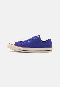 Des baskets basses en daim violet avec un capuchon en caoutchouc blanc à l'avant, des œillets blancs et une semelle intermédiaire crème avec une bande rouge et un accent bleu.