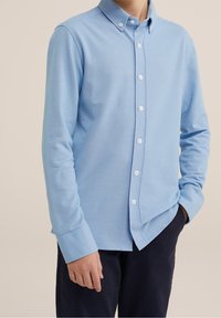 Světle modrá košile s dlouhým rukávem a knoflíkovým zapínáním vyrobená z texturované tkaniny, s límečkem a bílými knoflíky, doplněná tmavými kalhotami.
