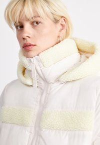 Kurtka puchowa w jasnym kremowym kolorze, z futrzanym żółtym kapturem i paskami na klatce piersiowej. Zamek błyskawiczny i gładka tekstura materiału.