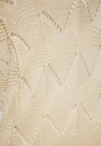 Țesătură tricotată de culoare bej deschis, cu un model texturat în zigzag și detalii de dantelă, creând un design decorativ și respirabil.