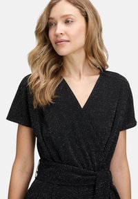 Robe noire à manches courtes, présentant un décolleté en V, un tissu texturé avec des paillettes argentées, et une ceinture nouée pour un effet cintré.