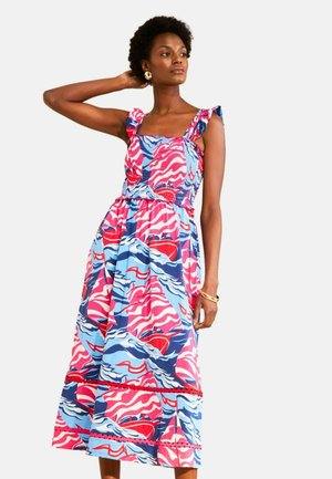 Kobieta ubrana w bezrękawnik, kolorową sukienkę midi w różowe, niebieskie i czerwone wzory z falbaniastymi ramiączkami, stojąca z jedną ręką uniesioną za głową.