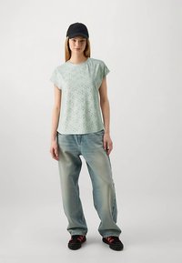 Lys grøn kortærmet top med et struktureret mønster, kombineret med falmede blå baggy jeans og sorte sko, suppleret med en sort kasket.