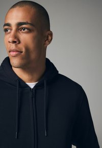 Haut à zip noir avec cordon de serrage, présentant une texture lisse. T-shirt blanc visible en dessous, sur un fond neutre.