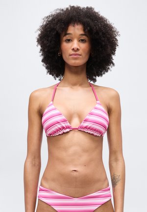 Žena s kučeravými vlasmi, oblečená v bikinách s ružovo-bielymi pruhmi, stojaca na jednoduchom svetlom pozadí.