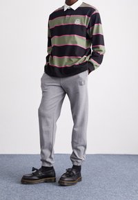 Person med långärmad svart och grönrandig skjorta med krage, grå mjukisbyxor och svarta skor med fransar, står på heltäckningsmatta.