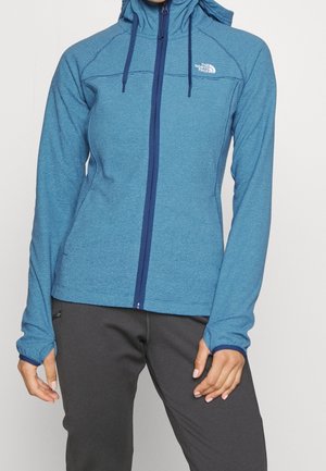 Sweat à capuche bleu zippé avec capuche et trous pour les pouces, porté avec un pantalon gris foncé, affichant le logo North Face sur le côté gauche de la poitrine.