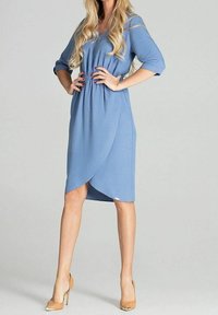 Eine Frau in einem blauen Wickelkleid und beigen High Heels steht selbstbewusst da, mit langen blonden Haaren, die über ihre Schultern fallen, vor einem hellen Hintergrund.