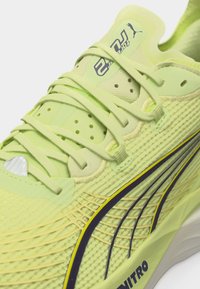 Κοντινό πλάνο ανοιχτό πράσινου παπουτσιού τρεξίματος Puma με κορδόνια, υφή από δίχτυ και τη λέξη «NITRO» τυπωμένη στη σόλα κοντά σε μαύρη και κίτρινη λωρίδα λογότυπου.