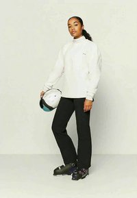 Donna che indossa un maglione bianco, pantaloni neri, scarponi da sci, mentre tiene un casco da sci bianco in mano, in piedi di fronte a uno sfondo chiaro e semplice.