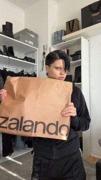 Pessoa com cabelo curto e escuro segurando uma grande sacola de compras da Zalando em uma sala com prateleiras de sapatos e bolsas.