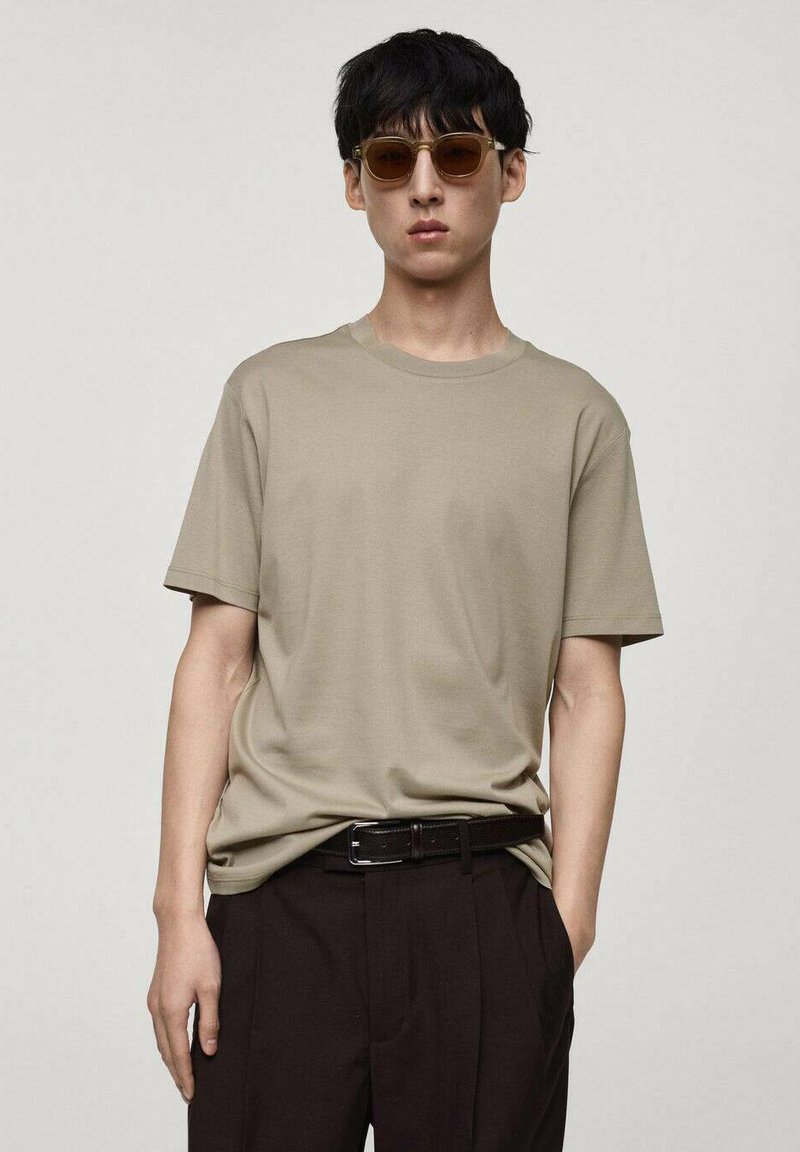 T-shirt in cotone beige con collo a giro, abbinato a pantaloni marroni e una cintura nera. Modello con occhiali da sole marroni.
