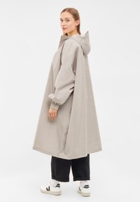 Abrigo impermeable gris claro con capucha, diseño oversize, puños elásticos y espalda sin costuras. Lleva puesto sobre pantalones negros y zapatillas blancas.