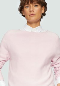 Helles rosa Sweatshirt mit strukturierter Oberfläche, entspannter Passform und Raglanärmeln. Über einem weißen Hemd mit Kragen getragen.