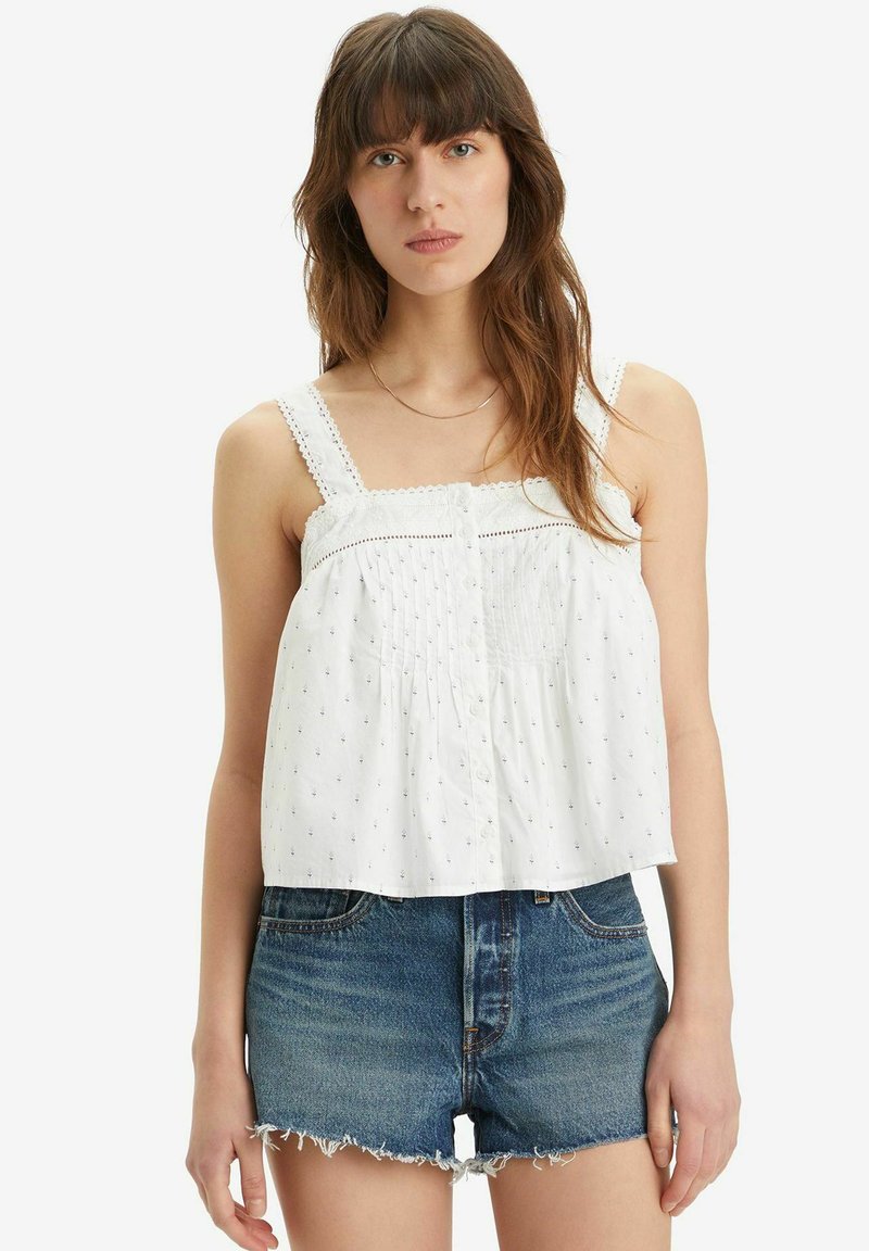 Hvid ærmeløs top med samlet front, dekorative knapper og blonde detaljer. Parret med slidte denimshorts.