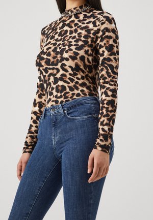 Kvinde iført en tætsiddende leopardmønstret langærmet top med høj hals, kombineret med blå højtaljede jeans og en sølvkæde halskæde.