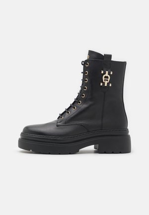 Bottine en cuir noir avec semelle épaisse, lacets à l'avant avec œillets dorés et boucle décorative dorée sur le côté.