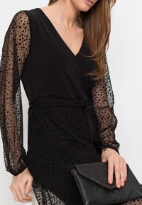 Svart klänning med texturerade prickiga genomskinliga ärmar, omslag design och en midjesnöre. Modellen håller en texturerad svart kuvertväska.