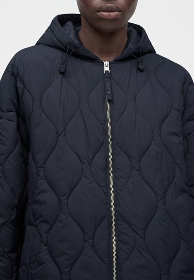 Personne portant une veste matelassée noire à capuche, avec une fermeture éclair frontale et des cordons à la capuche, visible de la poitrine aux lèvres.