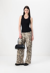 Haut noir sans manches avec des bordures en dentelle, associé à un pantalon large en imprimé serpent. Le mannequin tient un petit sac à main noir. Le sol est simplement blanc.