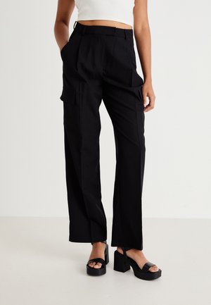 Pantaloni cargo neri con gambe larghe, due tasche laterali e una superficie liscia. Abbinati a sandali neri con plateau spessi.