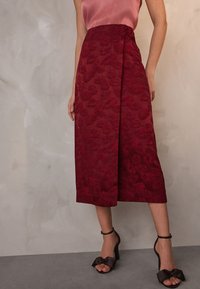 Piros virágmintás szoknya texturált felülettel és középmély hosszúsággal, fekete pántos magassarkú cipőkkel, melyeken masni díszítés található.