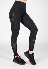 Zwarte sportlegging met een hoge taille, netaccenten bij de onderbenen en reflecterende branding. Gecombineerd met zwarte en rode sneakers.