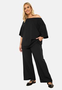 Svarta vida byxor i kombination med en oversized svart off-the-shoulder topp i mjukt tyg, som har en avslappnad passform och fickor.