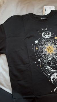Camiseta negra con un diseño solar y lunar que incluye elementos dorados, blancos y grises, representando planetas y estrellas sobre un fondo oscuro.