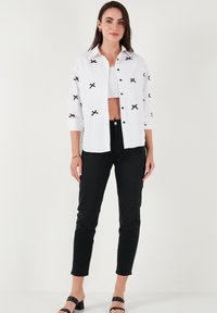White button-up shirt with black cross embroidery, paired with high-waisted black pants. Features a cropped white tank underneath.