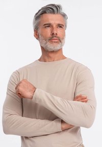 Ljus beige långärmad t-shirt gjord av mjukt tyg, med rund halsringning och åtsittande ärmar, bärs av en man med korslagda armar.