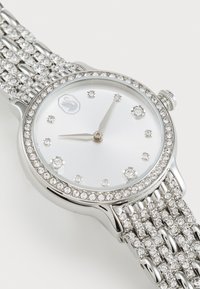 Silver-tone wristwatch with a circular face. White dial features crystal hour markers and a bezel set with stones. Multi-link bracelet design.