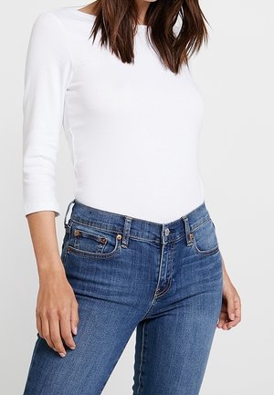 Frau trägt ein eng anliegendes weißes langärmliges Oberteil und blaue hoch taillierte Jeans, steht mit entspannt an den Seiten hängenden Händen vor hellem Hintergrund.