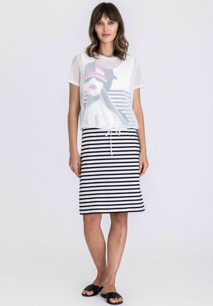 Femme aux cheveux bruns ondulés portant un t-shirt blanc à motif, une jupe rayée noir et blanc jusqu'aux genoux, et des sandales noires sans lacets, debout devant un fond blanc.