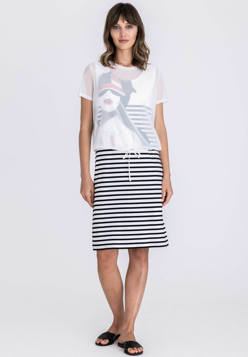 Vrouw met golvend bruin haar, gekleed in een wit T-shirt met afbeelding, een zwart-witte gestreepte knielange rok en zwarte instapsandalen, staand tegen een witte achtergrond.