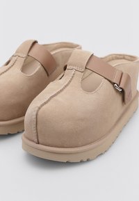 Zoccoli in suede beige con una fascia color cammello, dotati di un taglio anteriore, superficie testurizzata e suola in gomma spessa. Il design consente di indossarli facilmente.