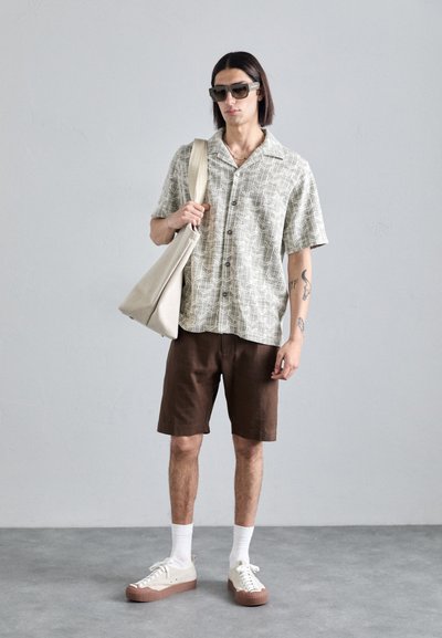 Hemd met korte mouwen in lichtbeige met een geometrisch patroon, bruine shorts en crèmekleurige sneakers met een bruine zool. Inclusief een canvas schoudertas.