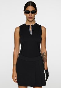 Svart golfklänning med hög halsringning, dragkedjeslutning och märkesdetaljer på kragen. Har en plisserad kjol och strukturerat material.