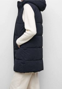 Černá prošívaná vesta s kapucí, na přední straně se zipem, postranními kapsami a prošívaným vzorem. Nošená přes světle béžový svetr.